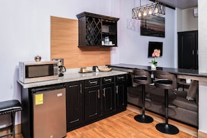 A compact kitchen with a microwave, coffee maker, and a wine rack. There are black cabinets, a wooden accent wall, and a bar stool area. - Shelbees Suites (Cornelia Ida)