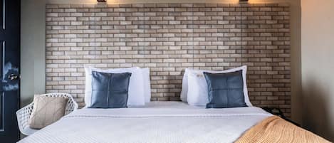 Ett hotellrum med en sänggavel som har ett mönster av tegelstenar, en säng med vita sängkläder, två kuddar, en vit stol och två vägglampor.