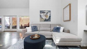 A modern living room with a sectional sofa, a round ottoman, and a coffee table. There is a framed abstract painting on the wall and a window with blinds. - The Polygon, Park Circle, Pet Friendly (North Charleston)