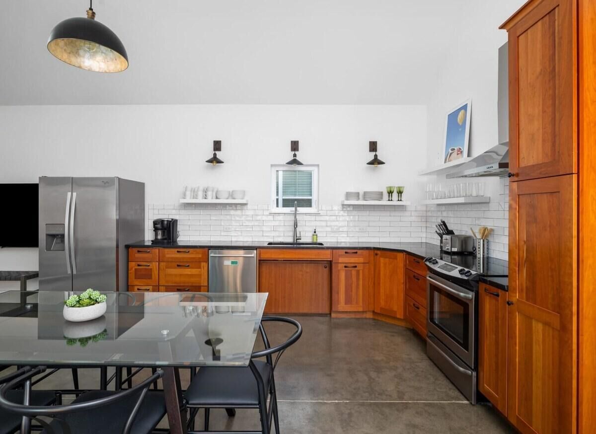 Een moderne keuken met een glazen eettafel, houten kastjes, roestvrijstalen apparatuur en hangende lampen.