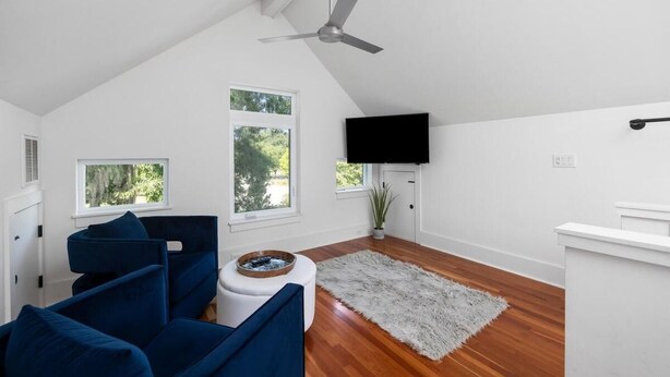 A modern living room with a sloped ceiling, a ceiling fan, a flat-screen TV, and a large window offering a view of greenery. - The Polygon, Park Circle, Pet Friendly (North Charleston)