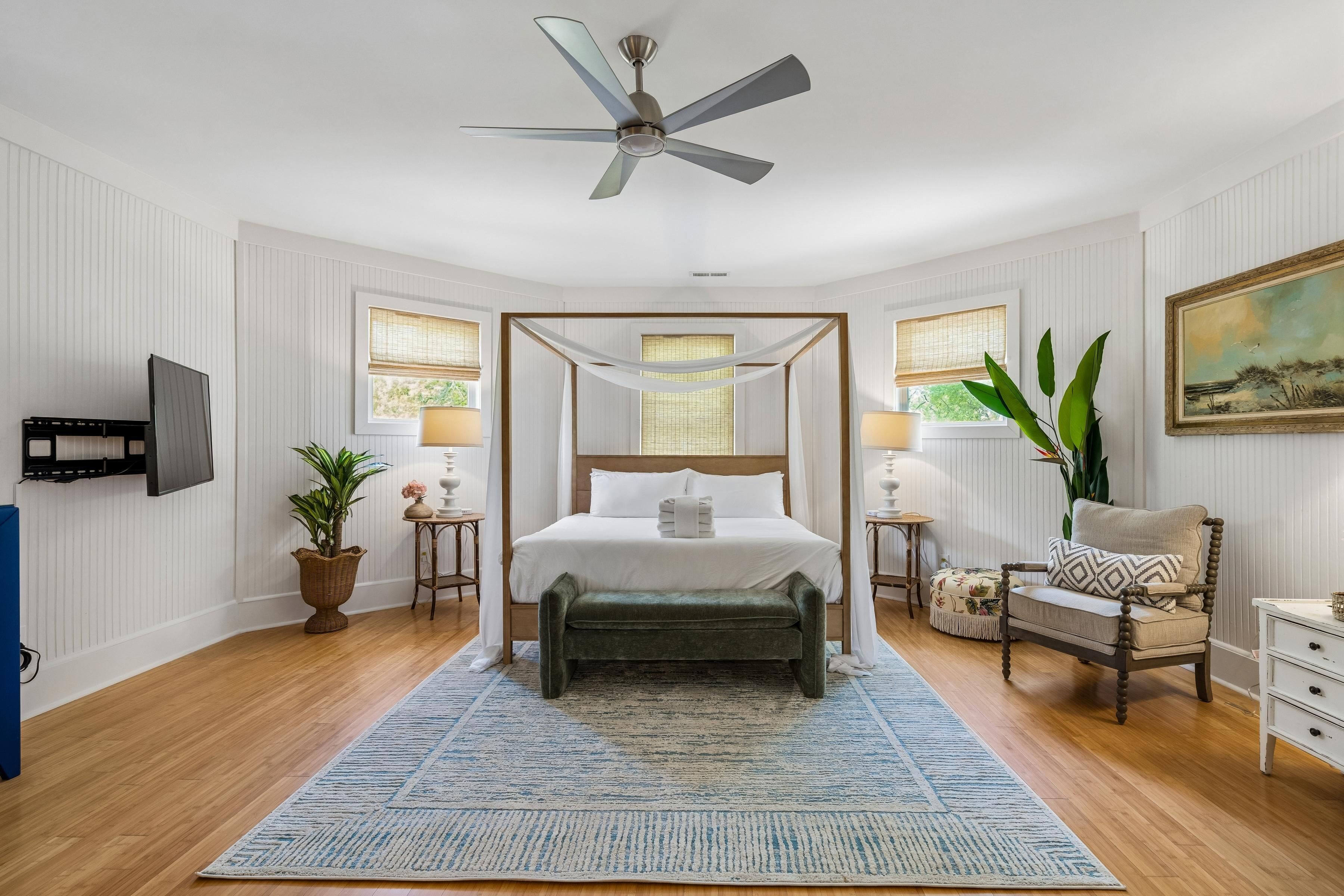 A bedroom with a four-poster bed, a ceiling fan, a TV mounted on the wall, and a large window with a view.