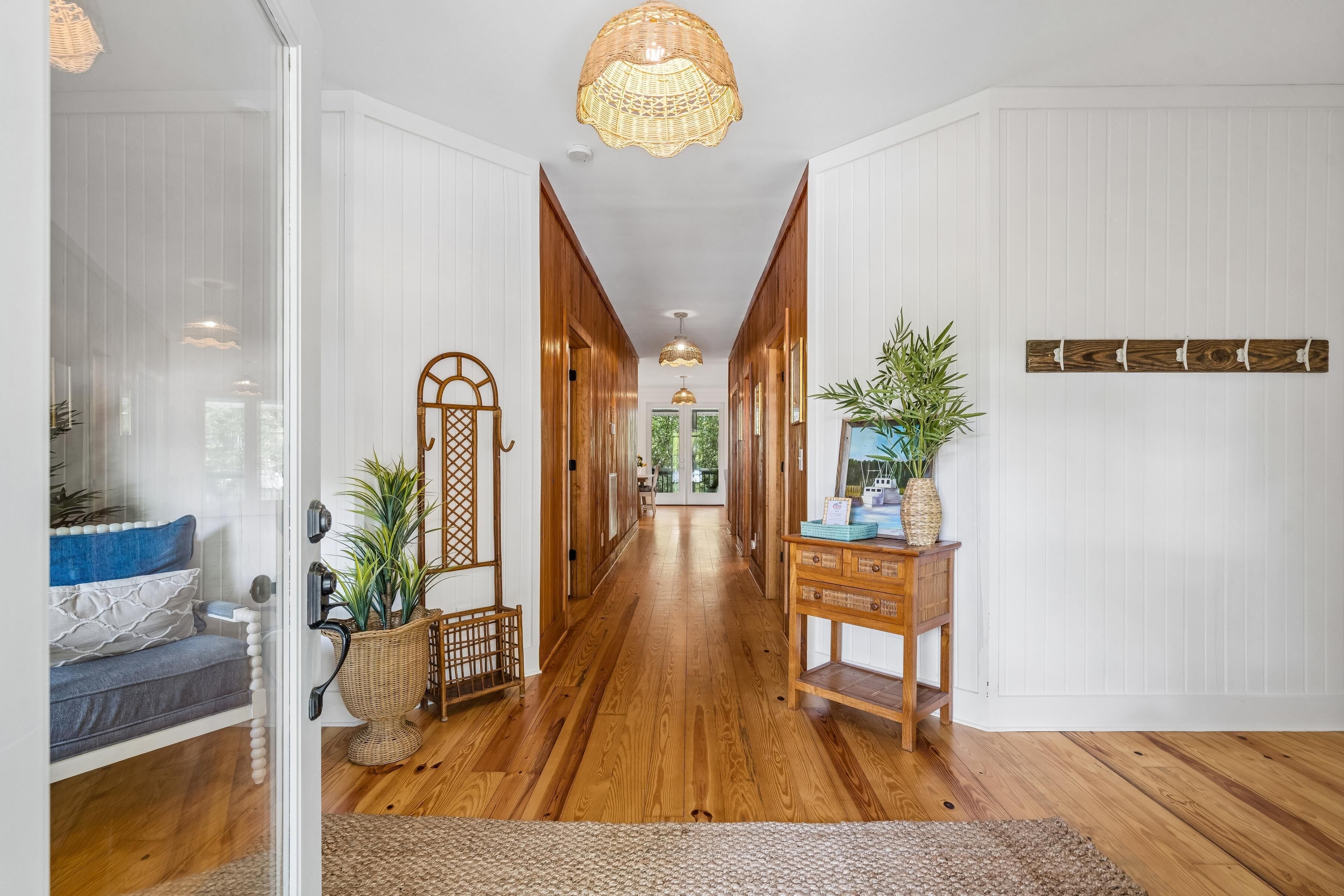 A hallway with wooden flooring, a wicker lamp, a wooden console table with plants, and a decorative wall piece.