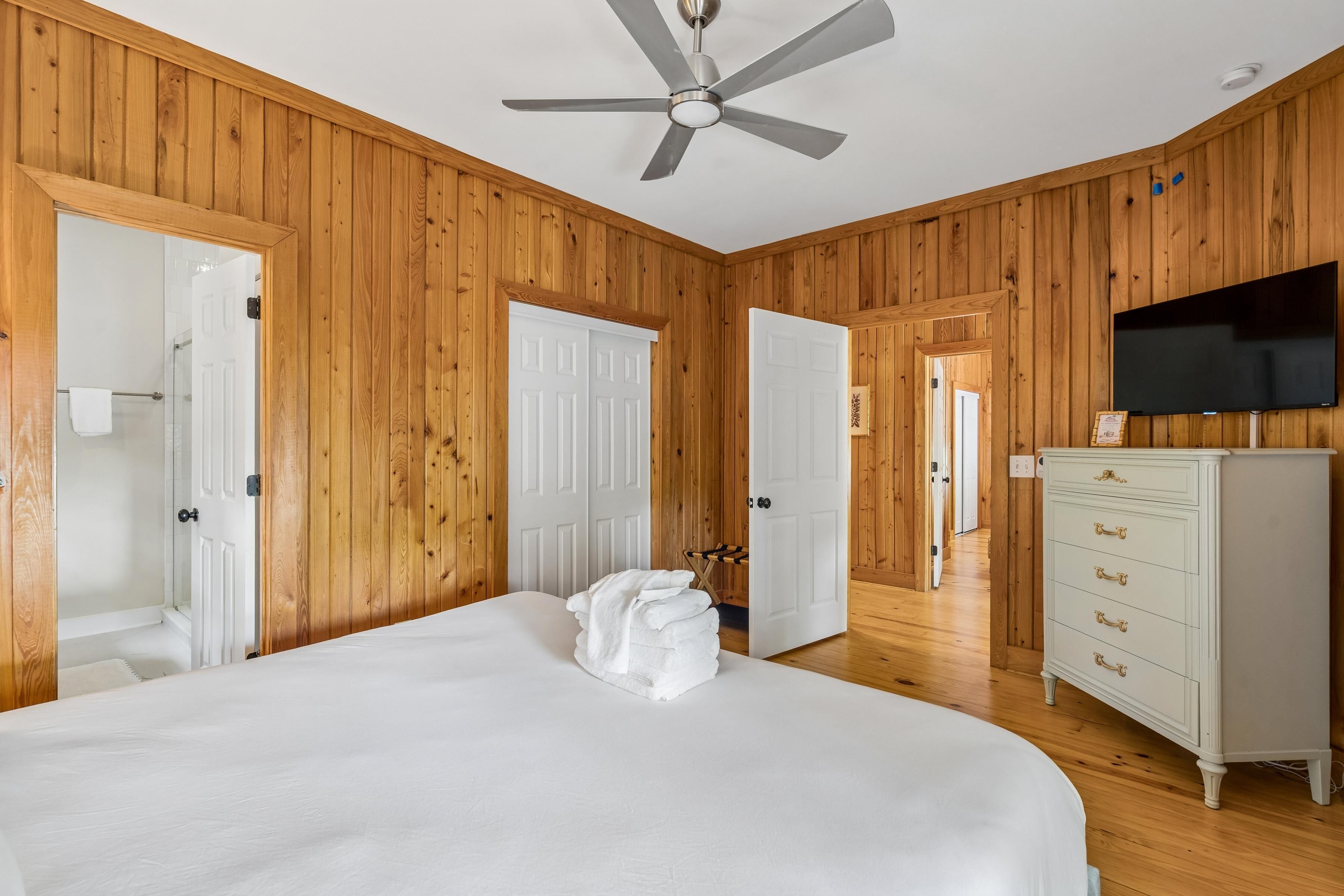 A room with wooden paneling, a bed with white linens, a ceiling fan, a TV on the wall, and a dresser.