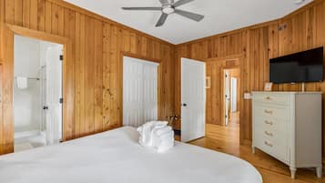 A room with wooden paneling, a bed with white linens, a ceiling fan, a TV on the wall, and a dresser.
