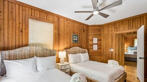 Una habitación con dos camas, un ventilador de techo, paredes de madera, una ventana con persianas y una puerta que da a otra habitación.