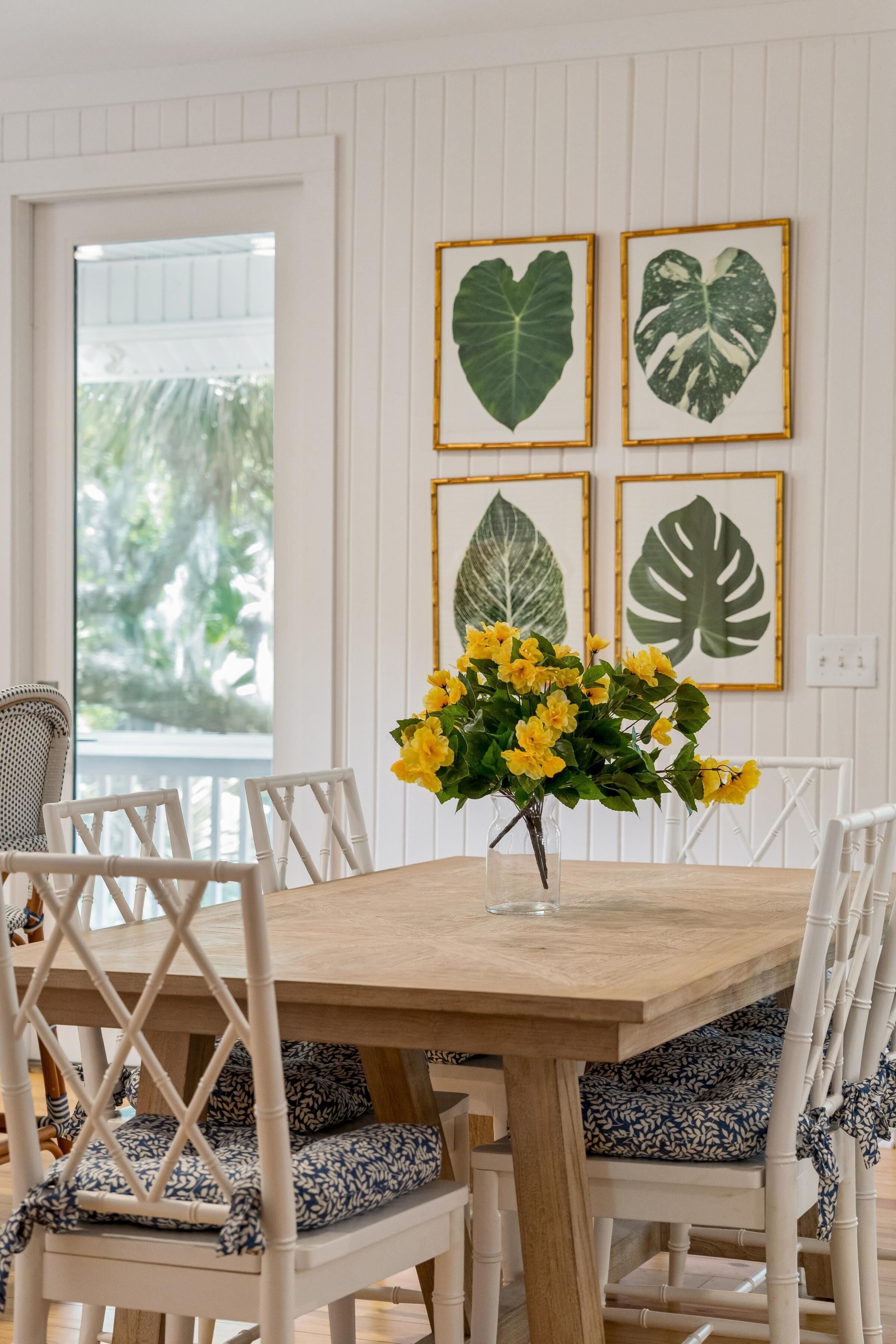 A dining table with chairs, a vase with yellow flowers, and framed leaf artwork on the wall.