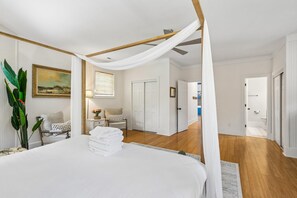 Quarto com cama, mesa de cabeceira, cadeira, planta e porta que dá para outro cômodo.