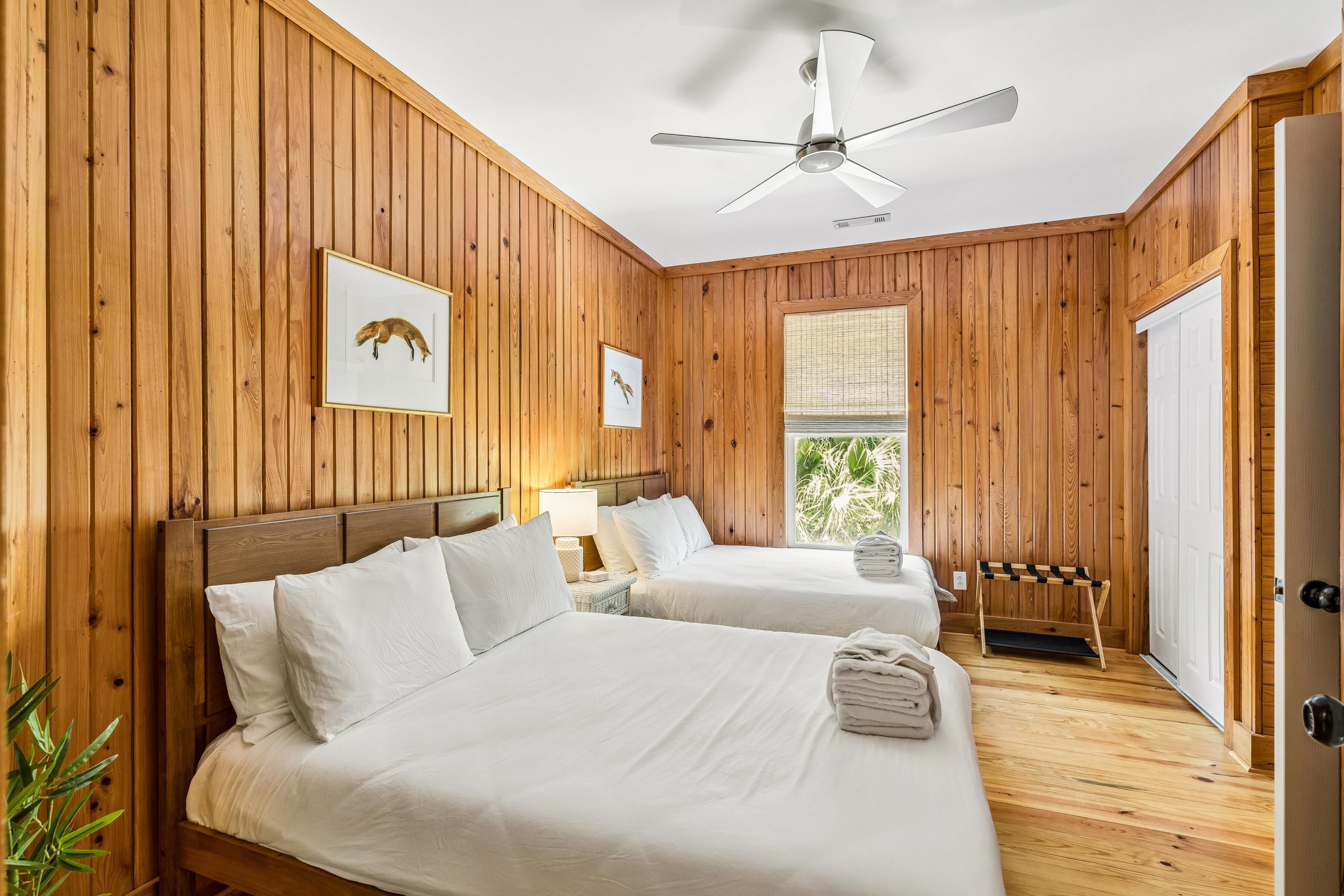 A room with wooden walls, two beds, a ceiling fan, and a window with a view.