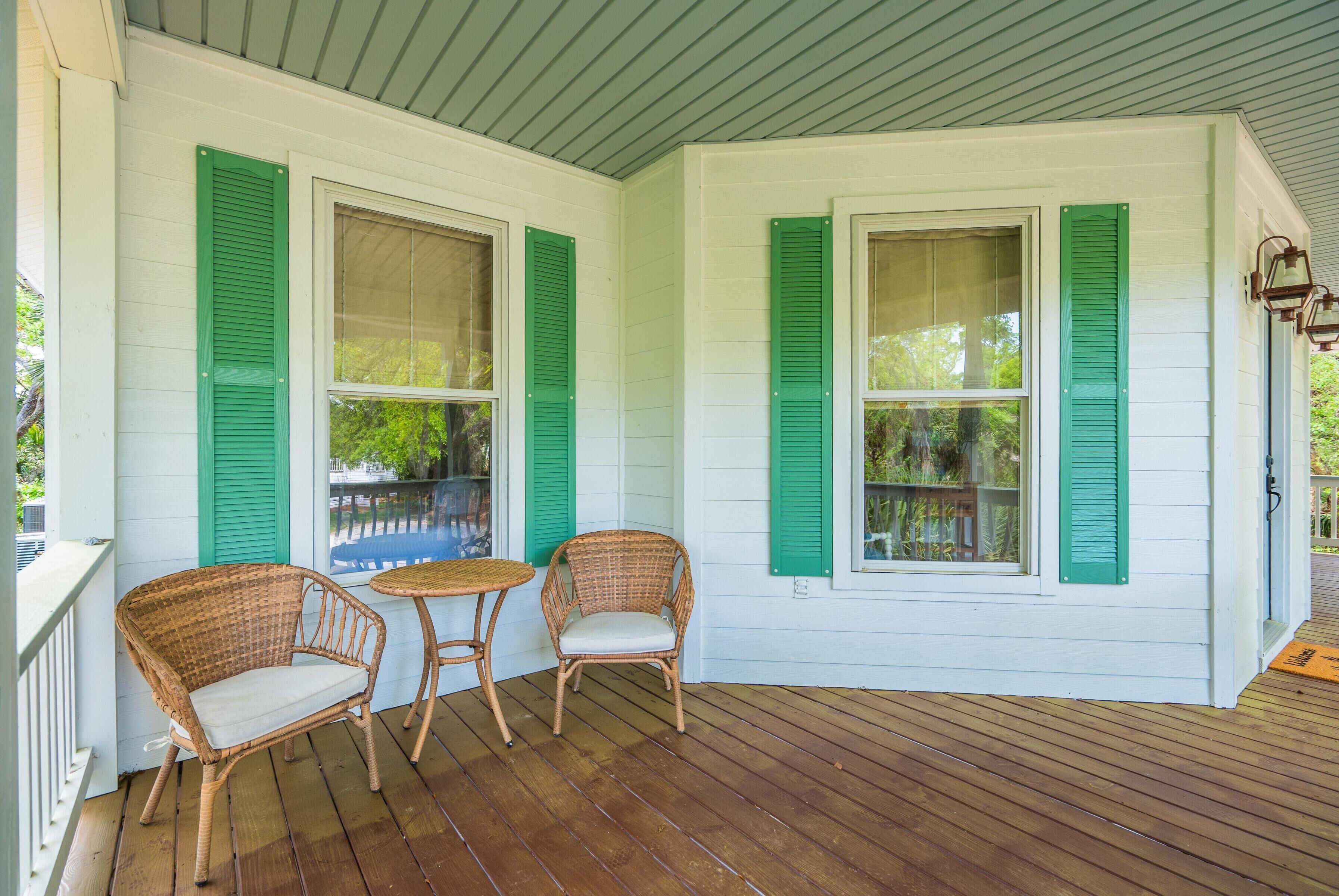 A covered porch with two wicker chairs and a small table, white exterior walls, and green shutters.
