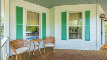 A covered porch with two wicker chairs and a small table, white exterior walls, and green shutters.