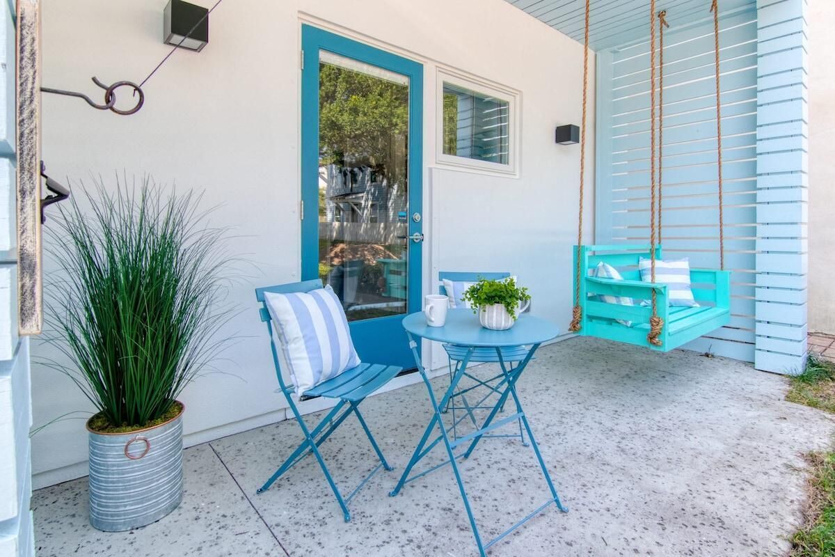 A small patio with a blue door, a swing chair, a potted plant, and a table with a pitcher and plant.