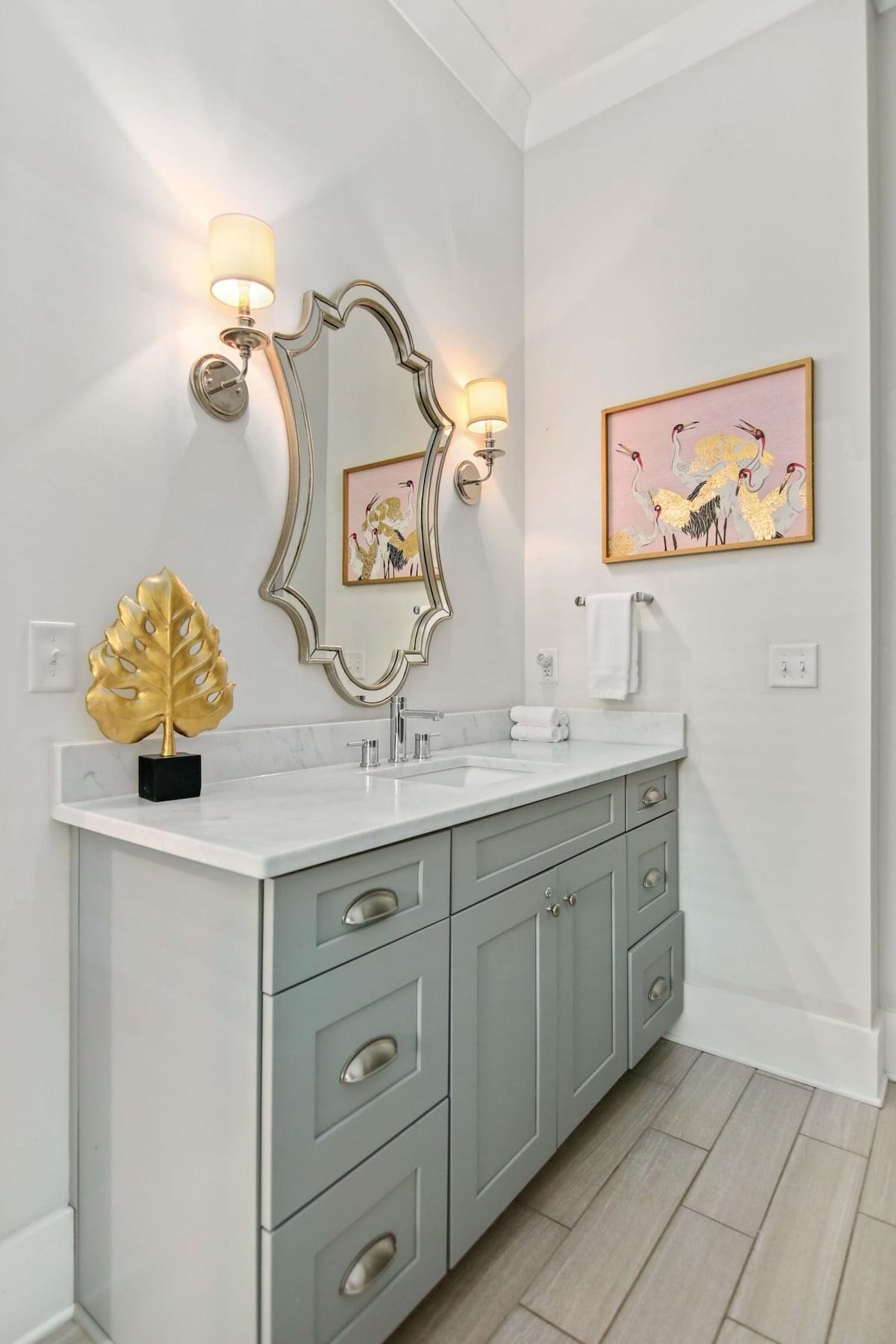 Une salle de bain moderne avec un meuble-lavabo blanc, des armoires grises, un motif décoratif en forme de feuille dorée et deux appliques murales.