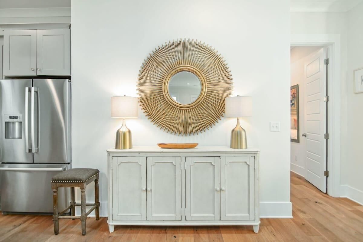 A kitchen with a white cabinet, a stainless steel refrigerator, a round mirror with a sunburst design, two lamps, and a wooden stool.