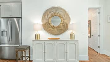A kitchen with a white cabinet, a stainless steel refrigerator, a round mirror with a sunburst design, two lamps, and a wooden stool.