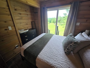 A wooden cabin bedroom with a bed, a dresser, and a view of a green landscape through a window. - Breeze & Blue Mountain Cabin:SLEEPS 12,Near CARRABASSETT/SUGARLOAF/HAS VIEWS (Eustis)