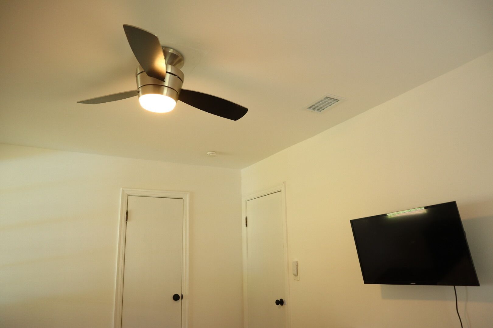 Een kamer met een plafondventilator, een aan de muur bevestigde televisie en twee gesloten deuren.