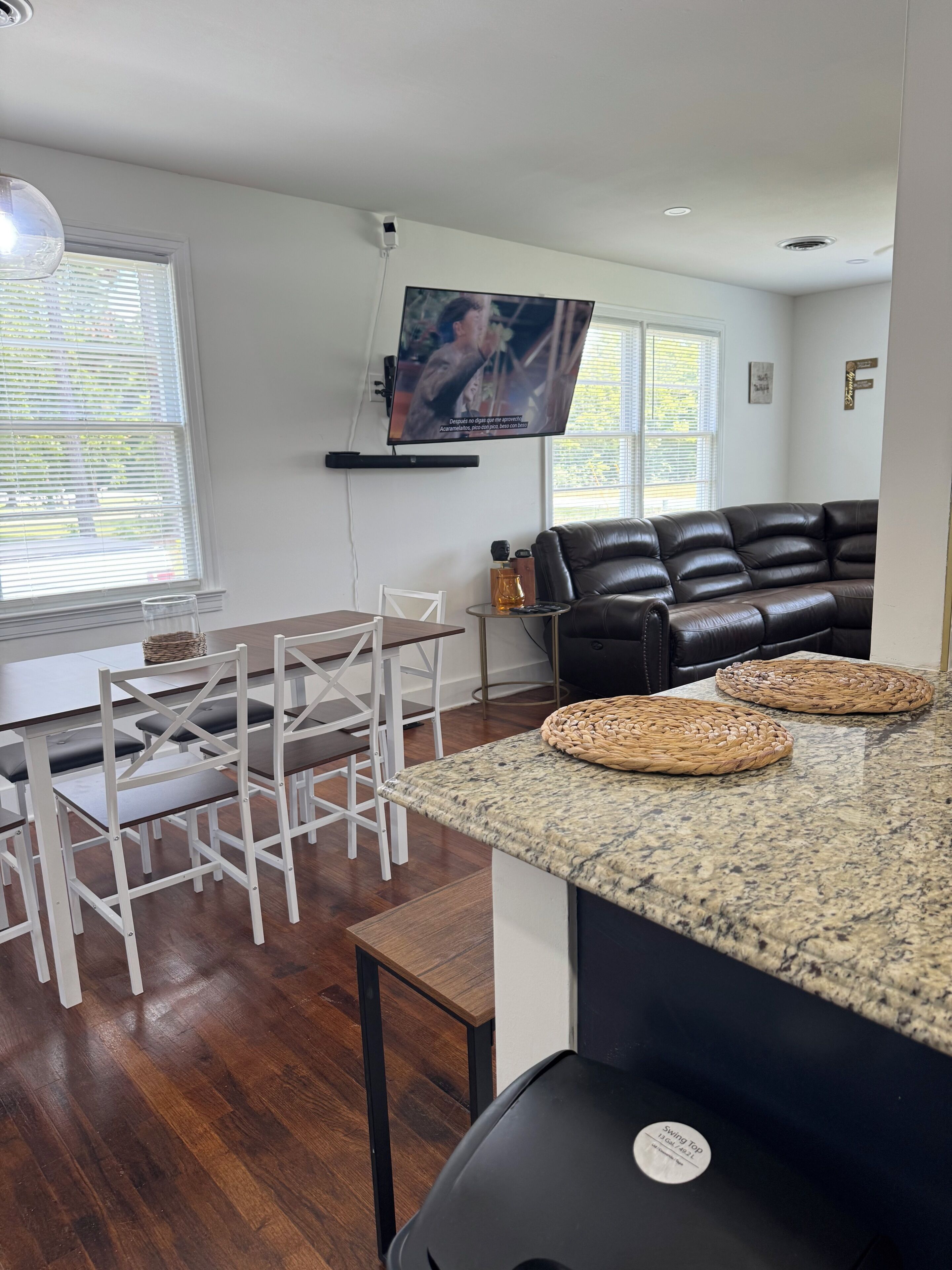 Una sala de estar moderna con un televisor de pantalla plana montado en la pared, un sofá de cuero negro, una zona de comedor con sillas blancas y una mesa de madera, y una encimera de cocina con individuales tejidos.