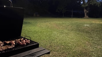 Un barbecue avec de la viande, placé sur une pelouse verdoyante, un banc en bois et des arbres en arrière-plan.