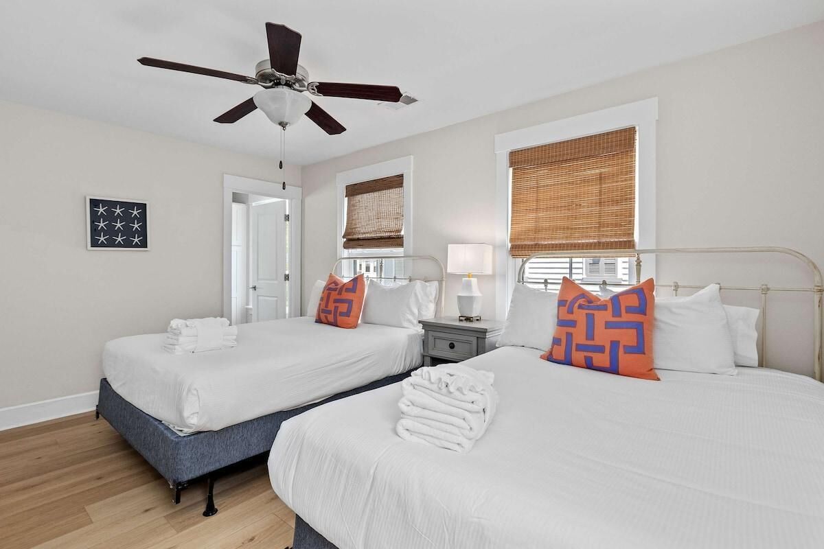 A hotel room with two beds, a ceiling fan, a window with blinds, a bedside table with a lamp, and a framed picture on the wall.