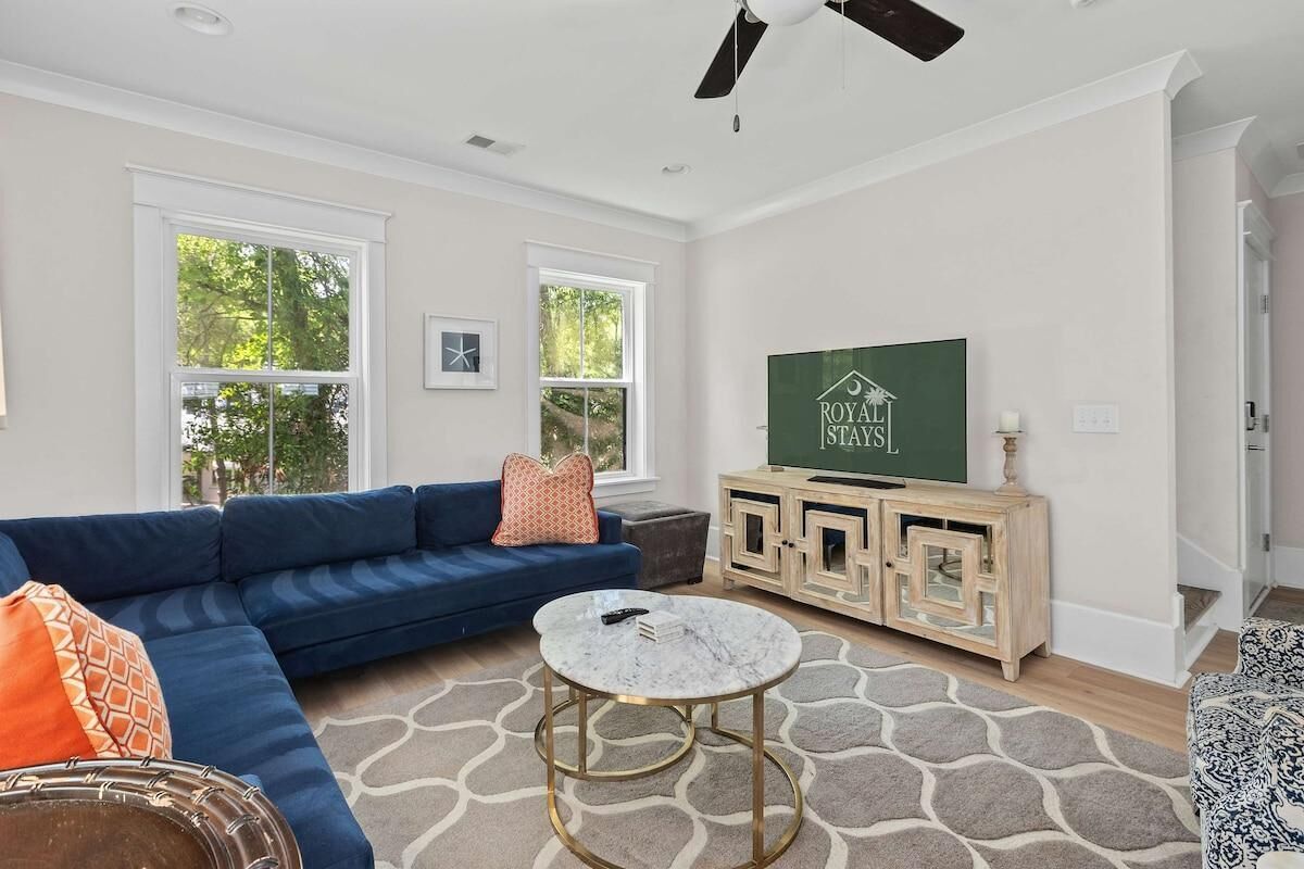 A living room with a blue sectional sofa, a round coffee table, a wooden cabinet with a TV, and large windows.