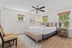 A bedroom with a bed, a chair, a bench, a nightstand, a ceiling fan, and windows with blinds. - Mt Pleasant Beach Retreat- Mins to Beach-hot Tub (Mount Pleasant)