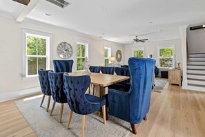 A dining area with a wooden table and blue upholstered chairs, large windows, and a ceiling fan. - Mt Pleasant Beach Retreat- Mins to Beach-hot Tub (Mount Pleasant)