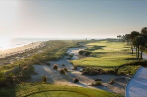 Un campo de golf costero con un sendero arenoso, fairways verdes y una playa al fondo.