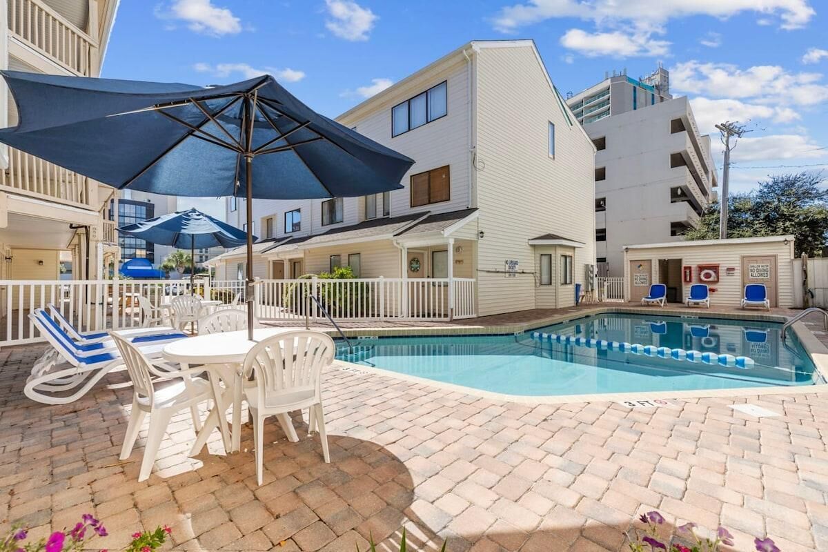 Un espace piscine aménagé avec des chaises longues et une table, entouré d’une clôture blanche, et jouxtant un bâtiment doté de nombreuses fenêtres et de balcons.
