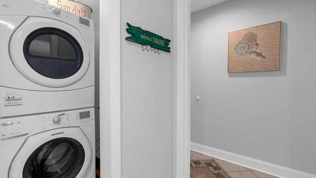 A laundry room with stacked washers and dryers, a green sign reading "Welcome to Paradise," and a framed artwork on the wall. - Magic Myrtle, On the Beach, Pet Friendly (Myrtle Beach)