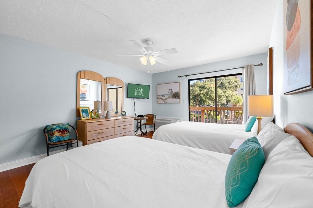 A hotel room with two beds, a desk, a chair, a ceiling fan, and a window with a view of the outdoors.