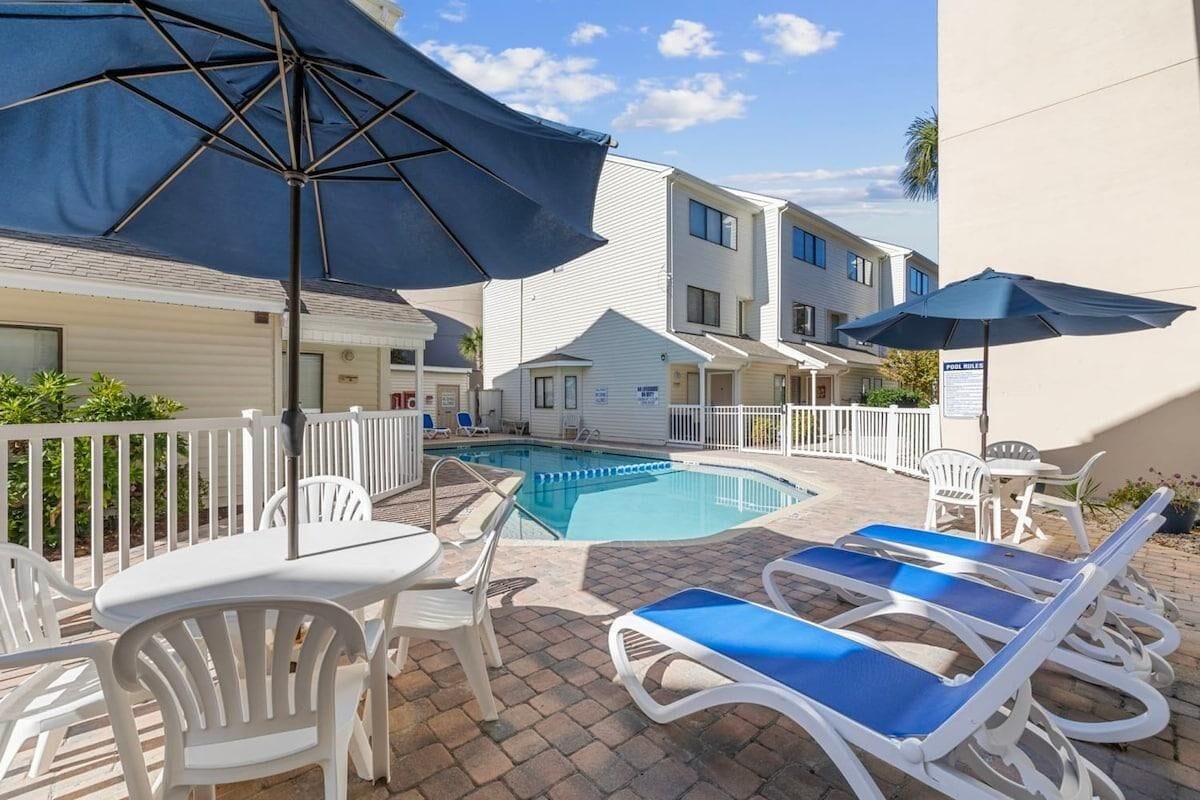 A pool area with lounge chairs and umbrellas, surrounded by a white fence and a building with multiple windows.