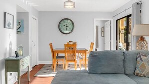 A living room with a grey sofa, a wooden dining table with chairs, a round wall clock, and a small side table. - Magic Myrtle, On the Beach, Pet Friendly (Myrtle Beach)