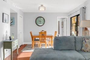 A living room with a grey sofa, a wooden dining table with chairs, a round wall clock, and a small side table. - Magic Myrtle, On the Beach, Pet Friendly (Myrtle Beach)