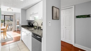 A modern kitchen with white cabinets, a granite countertop, and a dishwasher. There is a dining area with a table and chairs, and a view of the outside. - Magic Myrtle, On the Beach, Pet Friendly (Myrtle Beach)