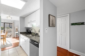 A modern kitchen with white cabinets, a granite countertop, and a dishwasher. There is a dining area with a table and chairs, and a view of the outside. - Magic Myrtle, On the Beach, Pet Friendly (Myrtle Beach)
