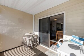 Uma área externa coberta com mesa e cadeiras, uma porta de vidro deslizante que dá para um espaço interno e um tapete estampado.