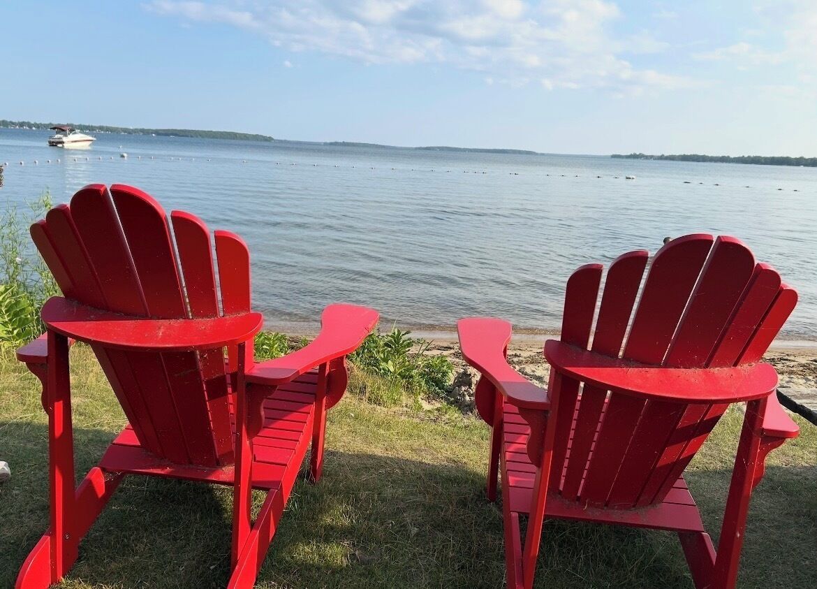 Deux chaises Adirondack rouges sont placées face à un lac paisible, avec un bateau visible au loin.