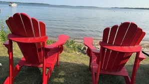 Two red Adirondack chairs facing a calm lake with a boat in the distance.