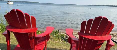 Two red Adirondack chairs facing a calm lake with a boat in the distance.