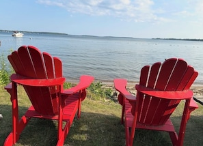 Deux chaises Adirondack rouges sont placées face à un lac paisible, avec un bateau visible au loin.