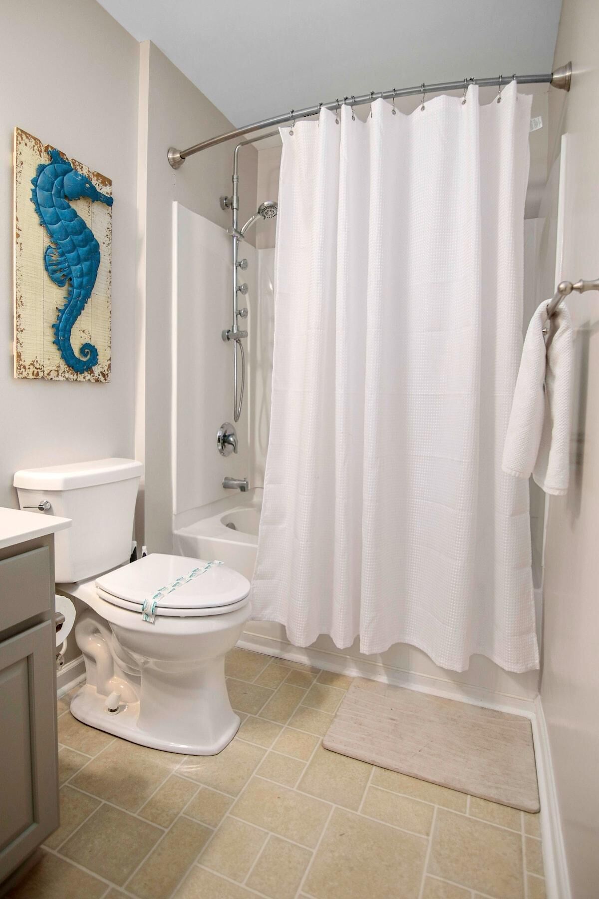 Een badkamer met een wit toilet, een bad met douchekop, een witte douchegordijn en een beige vloerkleed.