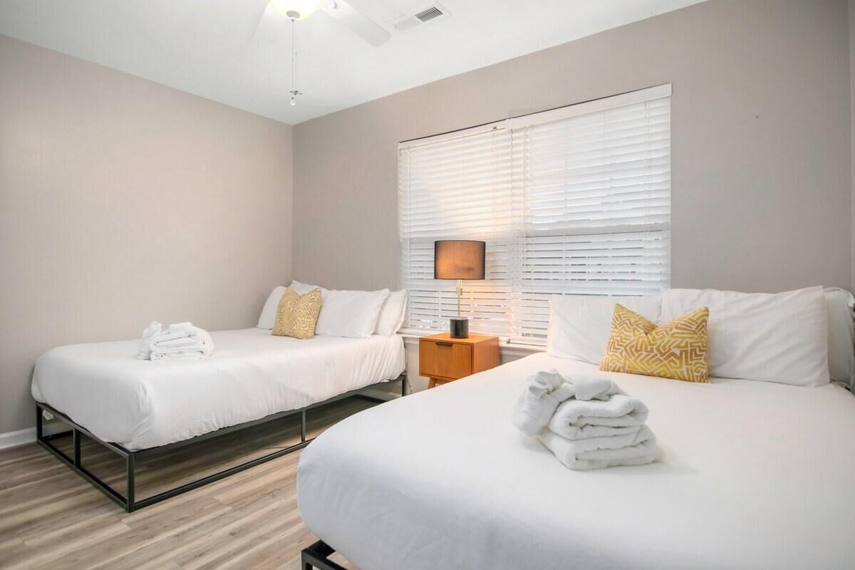 A hotel room with two beds, a ceiling fan, a nightstand with a lamp, and a window with blinds.