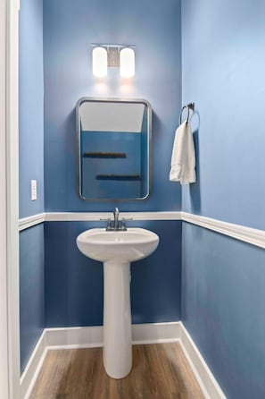 A bathroom with a white pedestal sink, a mirror with a light above it, and a towel hanging on a rack. - Penny Lane, Park Circle, 15m Dwtn CHS (North Charleston)