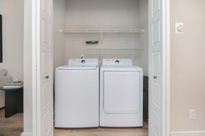 Un lavadero con dos lavadoras y secadoras de carga frontal blancas, un estante arriba y una puerta que conduce a otra habitación.