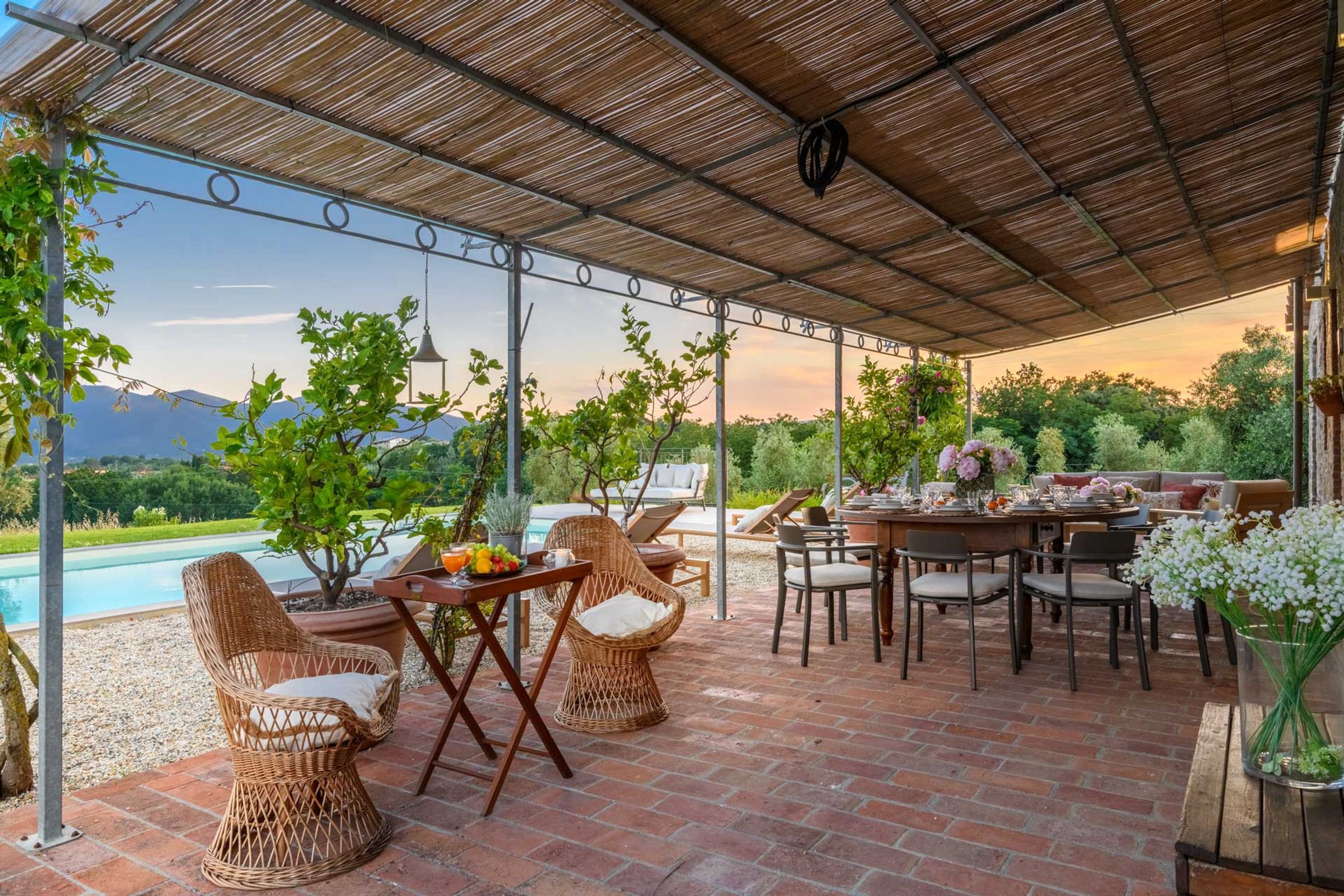 Un área de comedor al aire libre techada, con sillas de mimbre, una mesa preparada para una comida y vistas a una piscina y montañas.