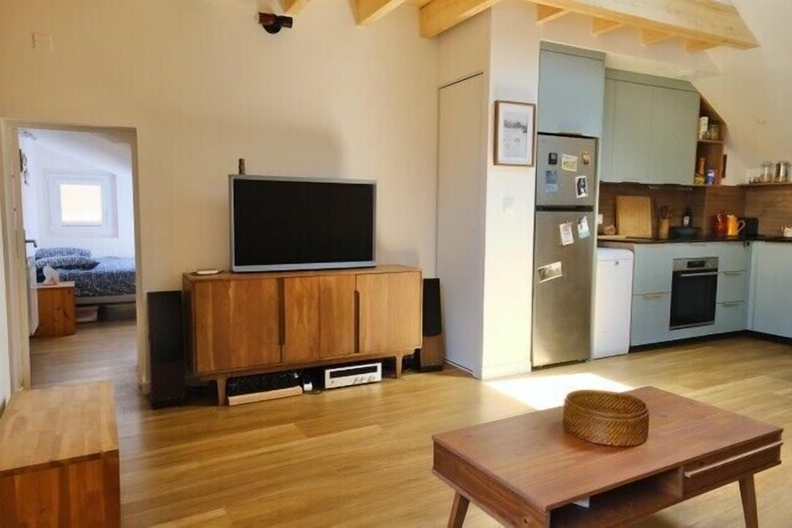 A living room with a television on a wooden cabinet, a dining table with a basket, and a kitchen area with cabinets and appliances.