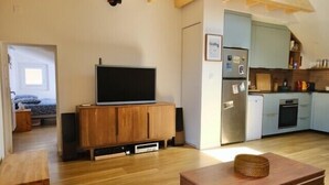 A living room with a television on a wooden cabinet, a dining table with a basket, and a kitchen area with cabinets and appliances. - Charming apartment in Rezé near Sèvre (Rezé)