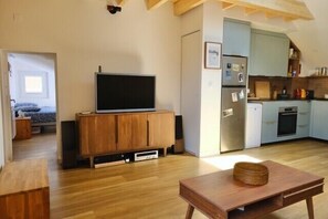 Ein Wohnzimmer mit Fernseher auf einer Holzkommode, einem Esstisch mit Korb und einer Küchenzeile mit Schränken und Geräten.