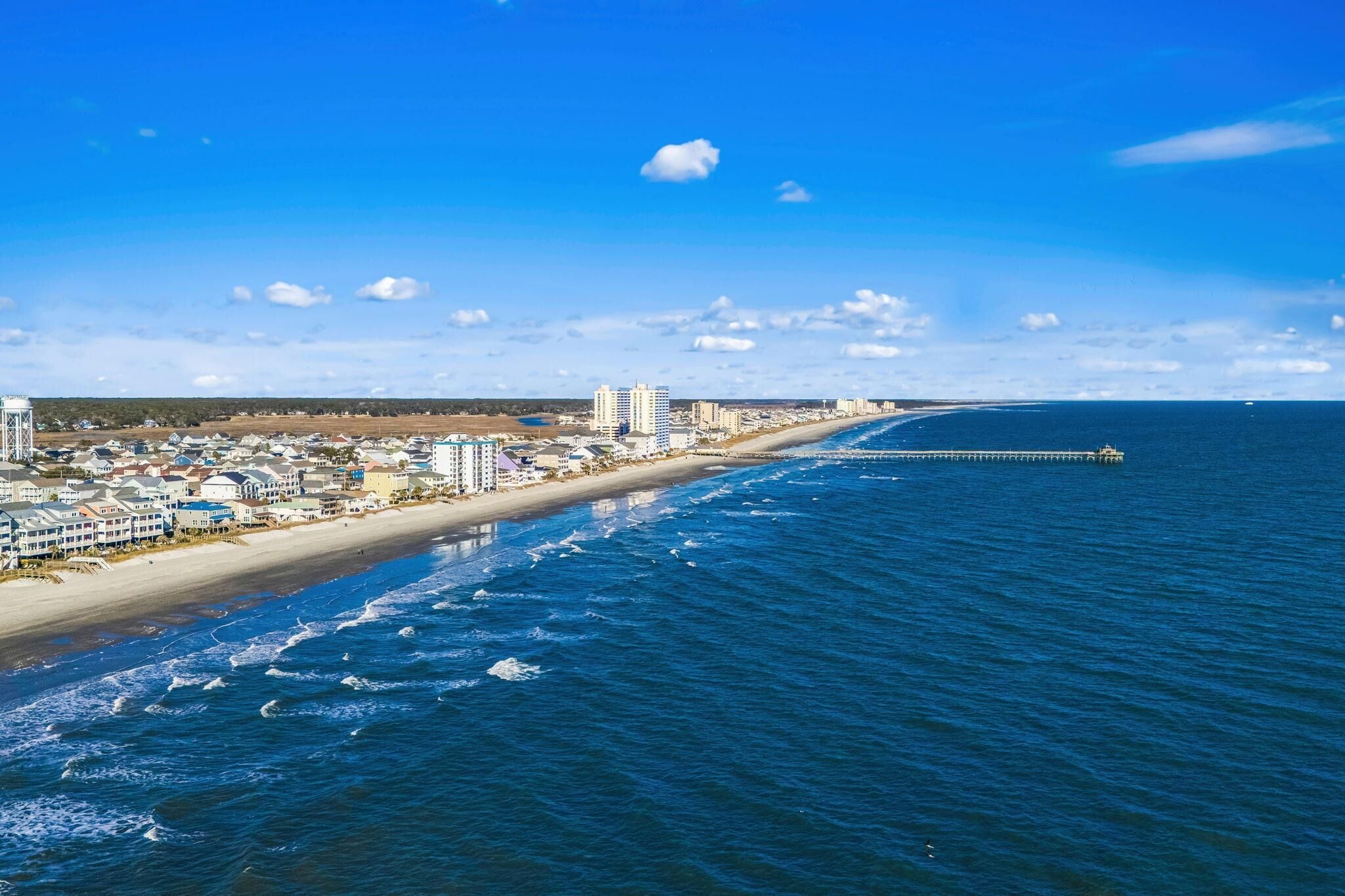 Un paisaje urbano costero con una playa, océano y un muelle que se adentra en el agua.